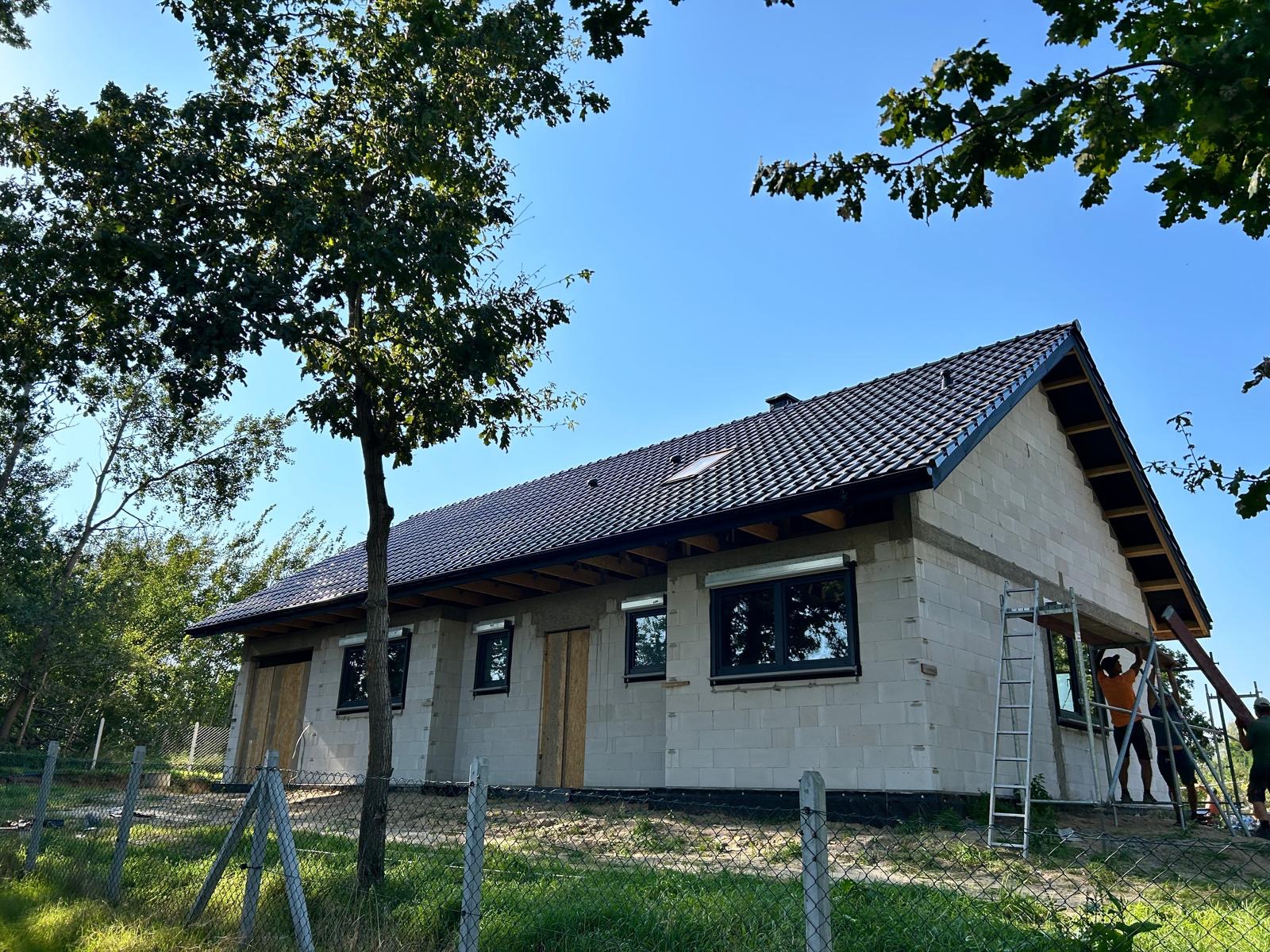 Dom w trakcie budowy z nowym, ciemnym dachem z dachówki ceramicznej i oknami z roletami zewnętrznymi, widoczne rusztowanie i pracownicy, ogrodzenie siatkowe na pierwszym planie.