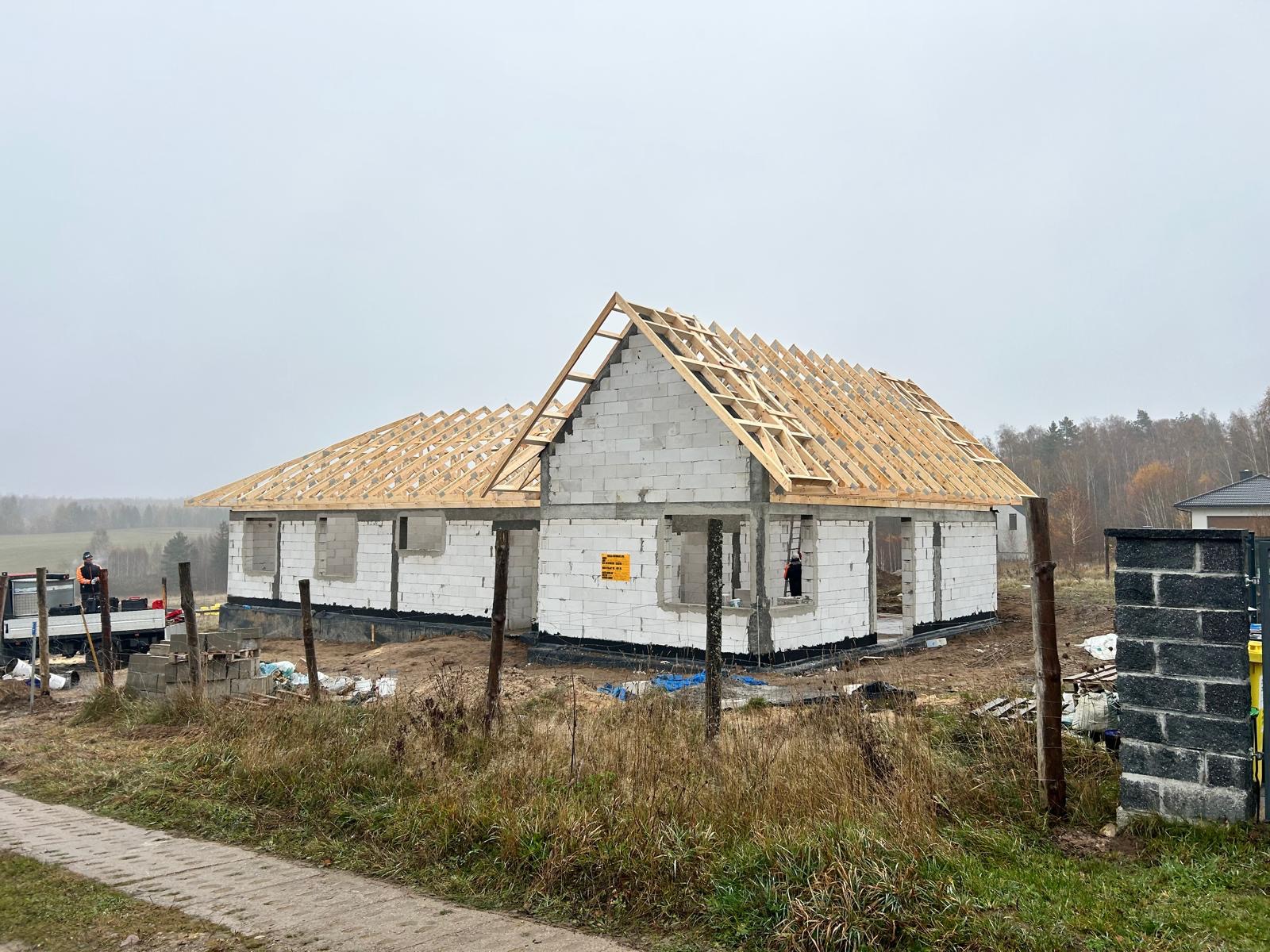 Surowy stan domu jednorodzinnego z białych bloczków, z widoczną drewnianą konstrukcją dachu w trakcie budowy, w tle krajobraz polny i pochmurne niebo.