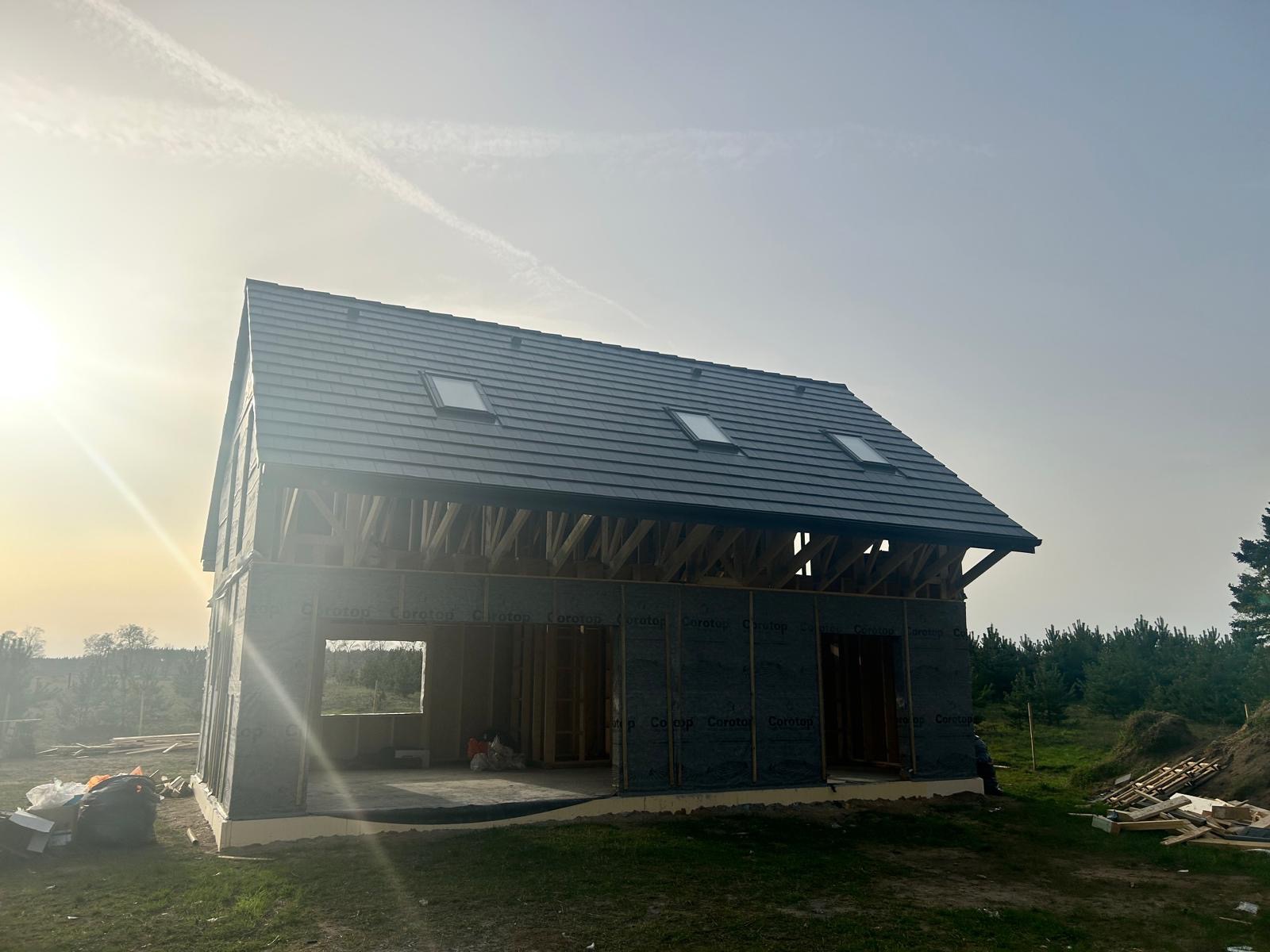 Wznoszony dom jednorodzinny z ciemną dachówką ceramiczną i trzema oknami dachowymi, widoczna konstrukcja więźby i membrana wiatroizolacyjna, w tle las i zachodzące słońce.