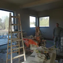 Wnętrze w trakcie prac wykończeniowych: drewniana drabina, maszyna do natrysku tynku PFT G4, worki z zaprawą, pracownik w tle.