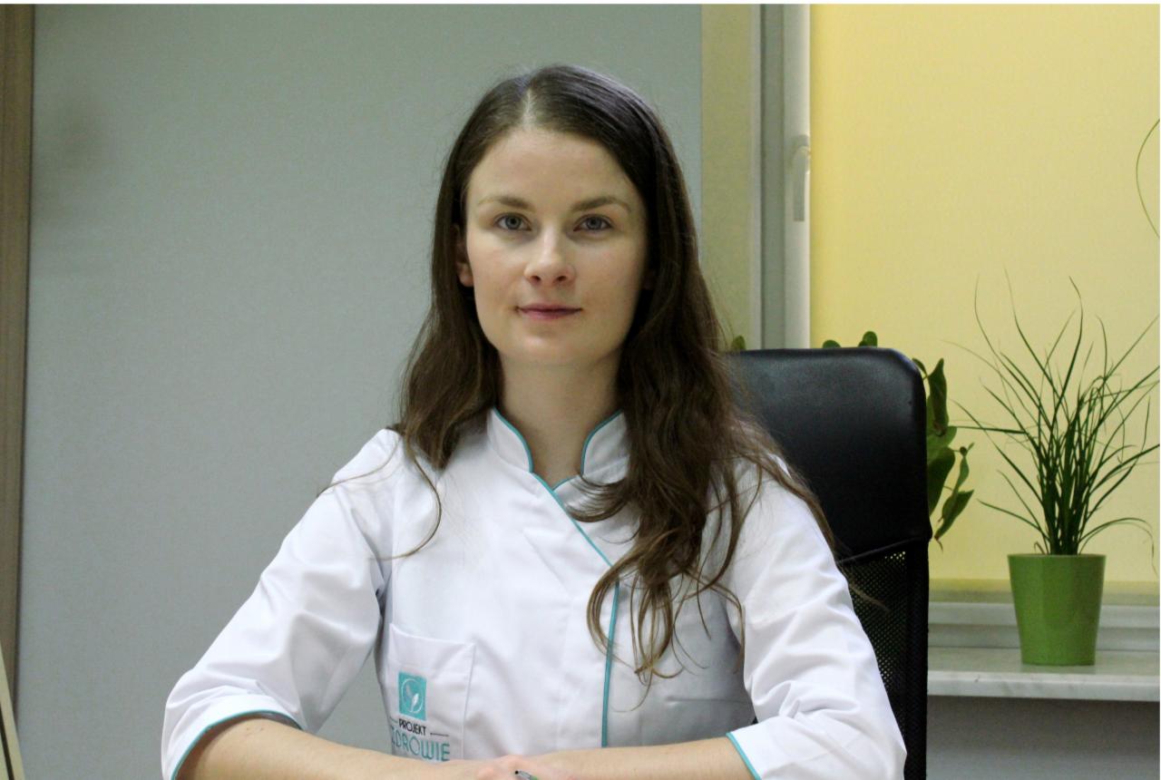 Portret kobiety w białym uniformie medycznym z logo 'Projekt Zdrowie' na tle biura z rośliną doniczkową.