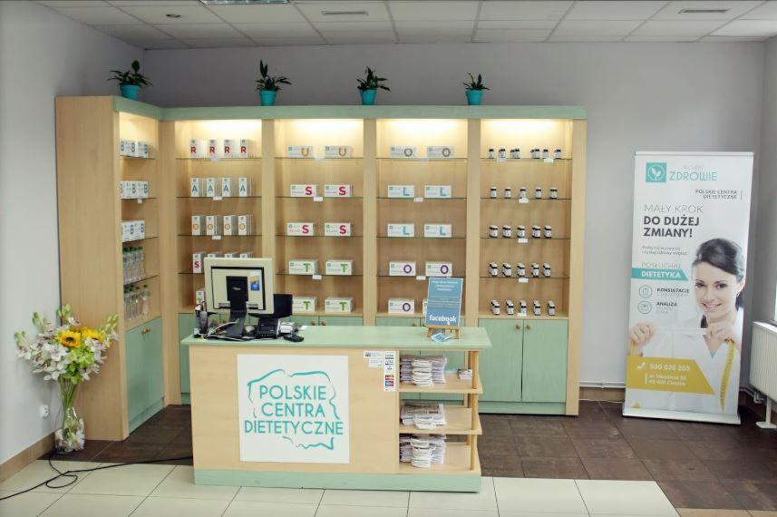 Wnętrze gabinetu dietetycznego: regały z suplementami, stanowisko obsługi klienta z logo 'Polskie Centra Dietetyczne' i baner reklamowy z uśmiechniętą dietetyczką.