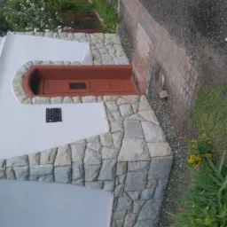 Wejście do budynku wykończone kamieniem, brązowe drzwi z małym okienkiem, tabliczka informacyjna na ścianie, obok ścieżka z kostki brukowej i trawa.