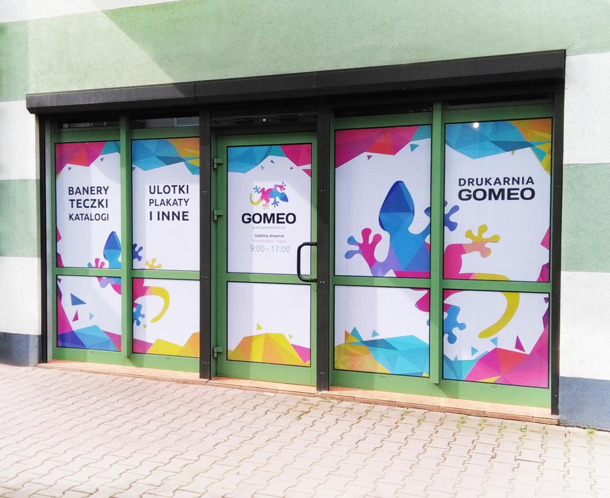 Fasada drukarni Gomeo z oklejonymi szybami, prezentującymi ofertę: banery, teczki, katalogi, plakaty, a także logo firmy z motywem kolorowego gekona w geometrycznym stylu.
