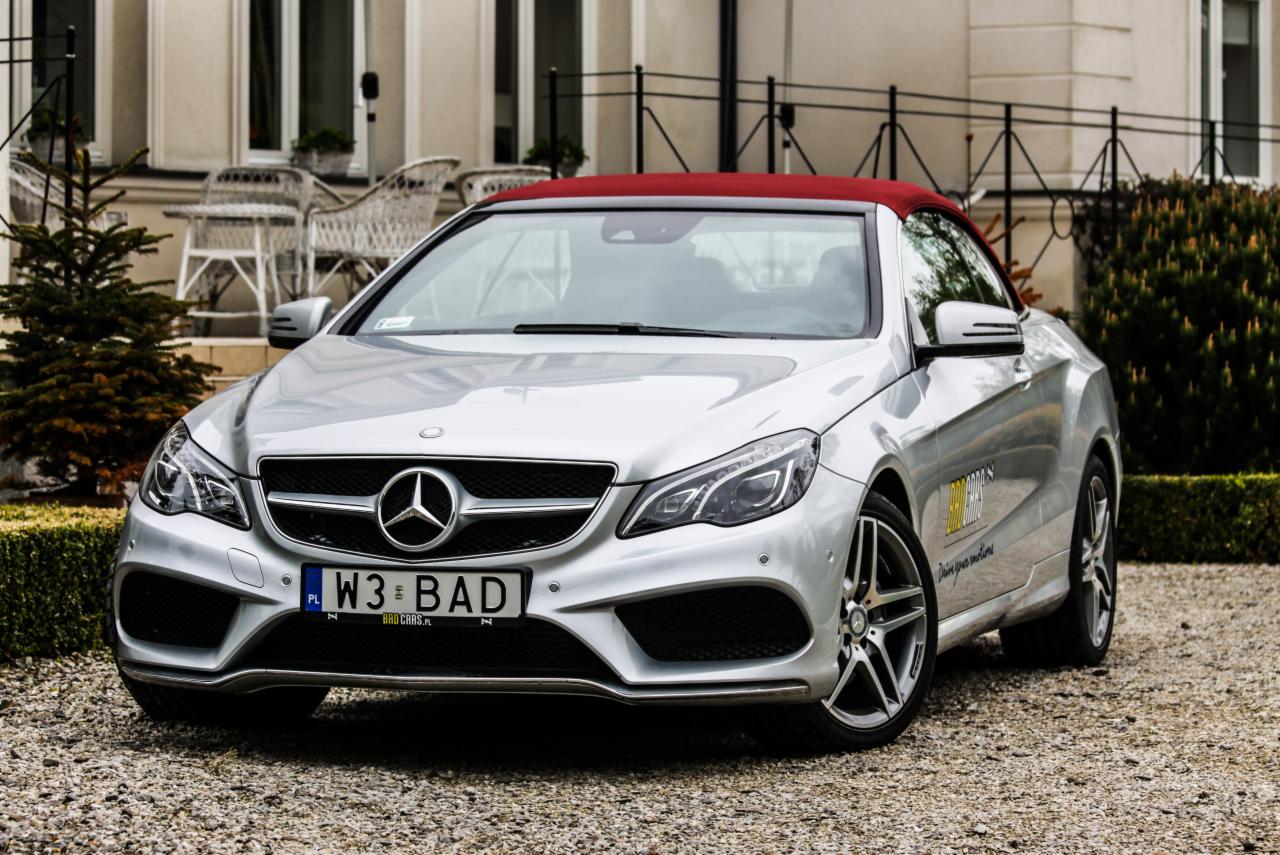 Srebrny kabriolet Mercedes-Benz z czerwonym dachem, zaparkowany na żwirowym podjeździe przed budynkiem z białymi kolumnami i rattanowymi krzesłami w tle.