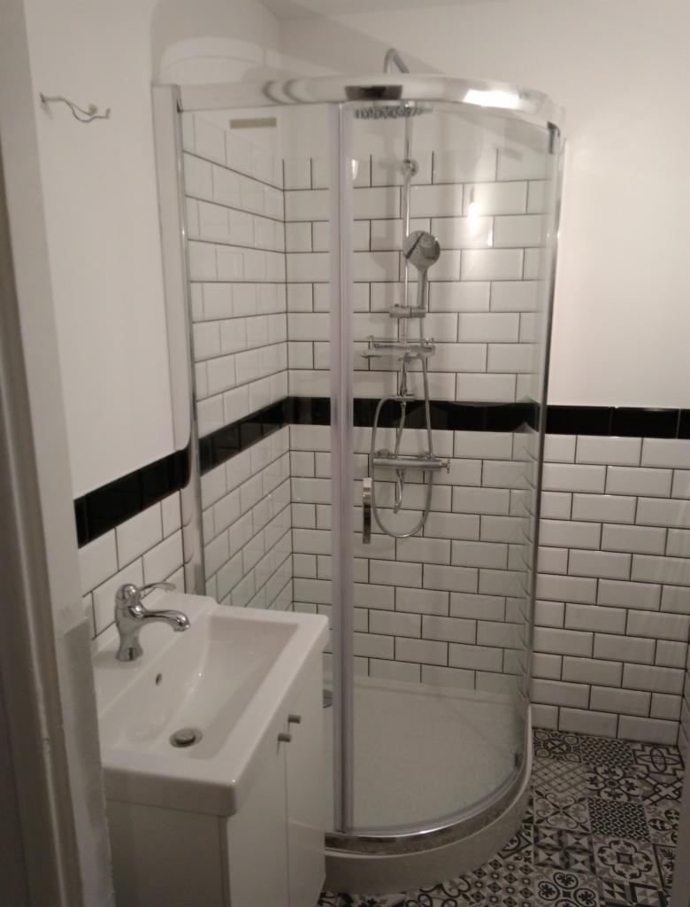 Nowoczesna łazienka z kabiną prysznicową narożną, białą umywalką z szafką i płytkami typu metro z czarnym pasem na ścianach oraz wzorzystą podłogą.