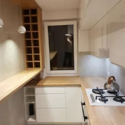 Jasna kuchnia z drewnianym blatem i białymi frontami szafek, widoczna płyta grzewcza i czajnik, na ścianie półki na wino, w oknie odbicie osoby.