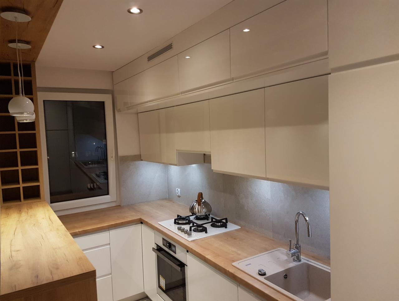 Nowoczesna kuchnia z białymi, lakierowanymi szafkami górnymi bez uchwytów, drewnianym blatem i płytą grzewczą, widok z perspektywy narożnika.