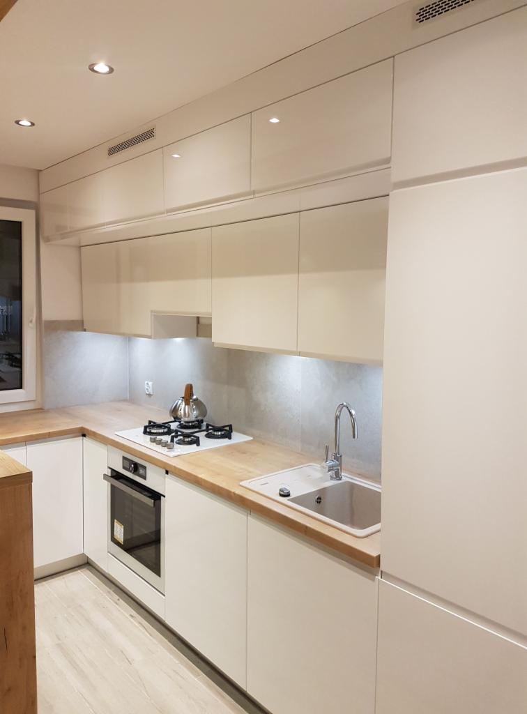 Nowoczesna kuchnia z białymi, lakierowanymi frontami, drewnianym blatem i płytą gazową z czajnikiem. Górne szafki z wbudowanym oświetleniem punktowym i kratką wentylacyjną.