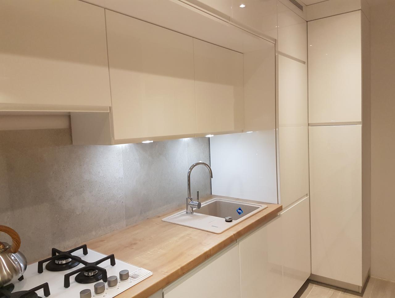 Jasna kuchnia z białymi, lakierowanymi szafkami, drewnianym blatem, ceramicznym zlewem i płytą gazową z czajnikiem na palniku.