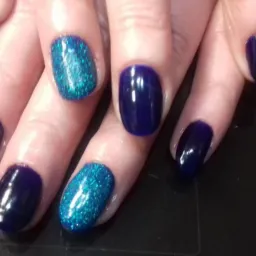 Dłoń z pomalowanymi paznokciami: ciemnogranatowe i turkusowe z brokatem, widoczna buteleczka lakieru w tle.