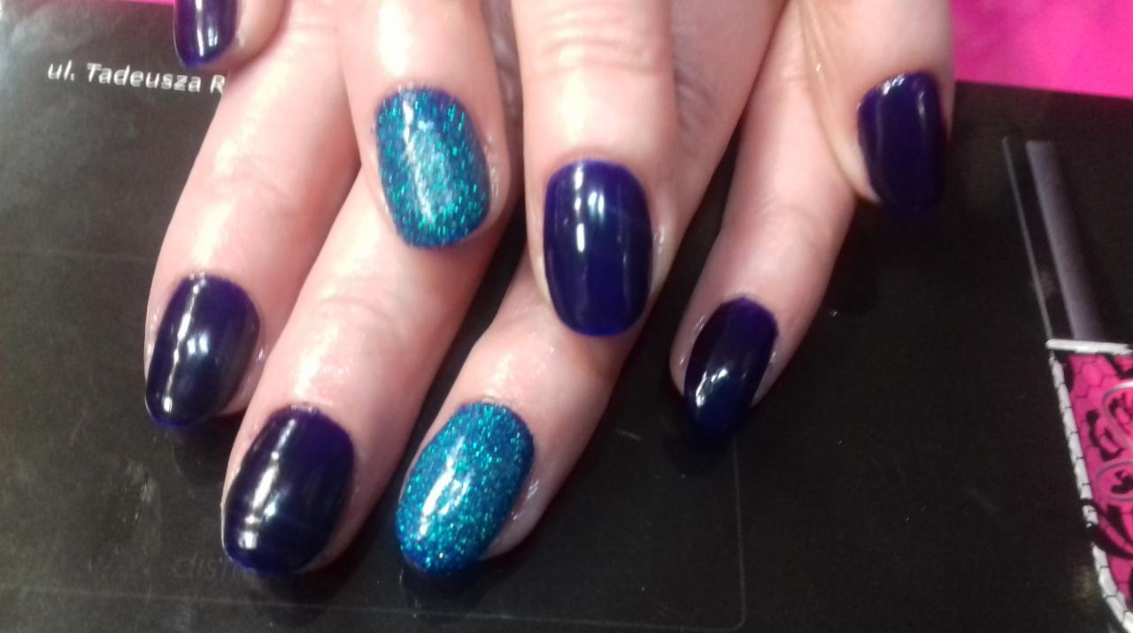 Dłoń z pomalowanymi paznokciami: ciemnogranatowe i turkusowe z brokatem, widoczna buteleczka lakieru w tle.