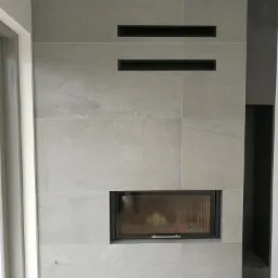 Obudowa kominka wykończona szarymi płytami kamiennymi, z czarnymi kratkami wentylacyjnymi i wkładem kominkowym, widoczna część podłogi z jasnego drewna.