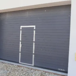 Szara brama garażowa segmentowa z drzwiami przejściowymi, widoczny fragment elewacji budynku i żółta skrzynka gazowa.