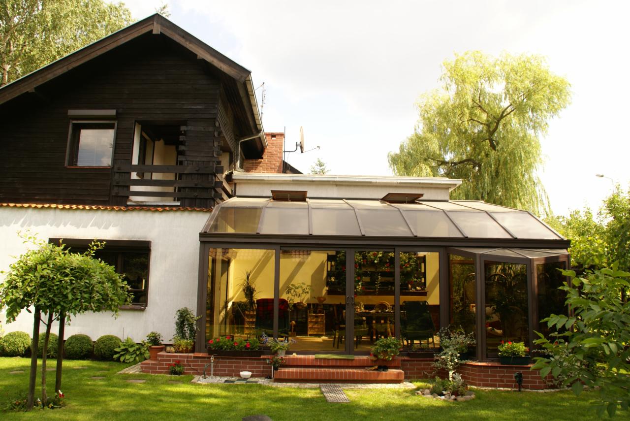 Przydomowa oranżeria z brązowymi ramami, dobudowana do dwukondygnacyjnego domu z drewnianą górą i białym dołem, widok z ogrodu.