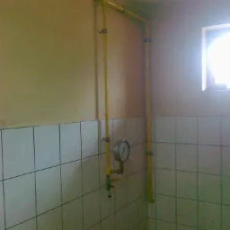 Żółta instalacja gazowa z manometrem na ścianie wyłożonej białymi płytkami, widok z dołu, naturalne światło z okna.