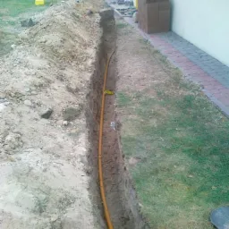 Żółty przewód w wykopie ziemnym wzdłuż ściany budynku, etap prac ziemnych przed zasypaniem.