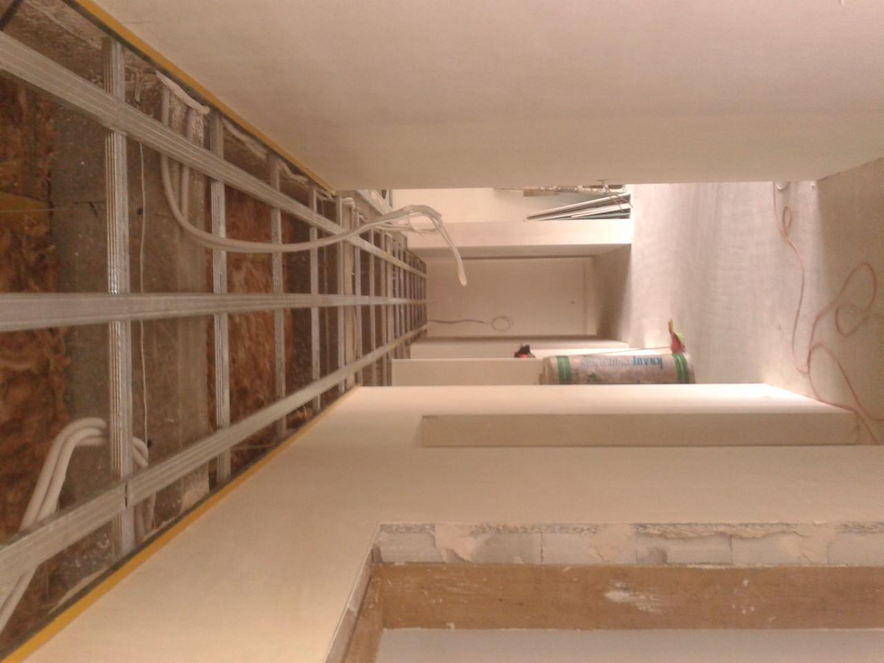 Wnętrze w trakcie prac wykończeniowych: metalowy stelaż z instalacją elektryczną, częściowo obłożony płytami, ściany przygotowane do malowania, widoczny wałek malarski i tuba masy szpachlowej.