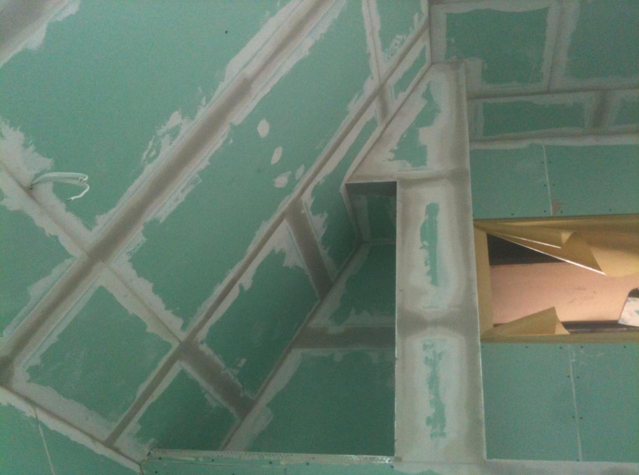 Skośny sufit i ściany pokryte zielonymi płytami, częściowo zaszpachlowane, widoczne kable i fragment okna zabezpieczonego folią malarską.