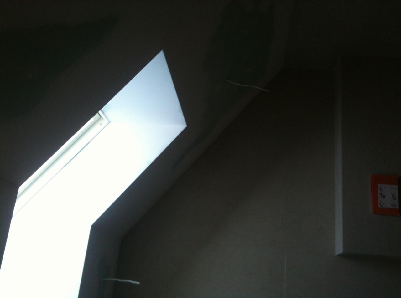 Wnętrze poddasza z oknem dachowym w trakcie wykańczania, widoczne niedoskonałości na ścianie i wystający przewód elektryczny.