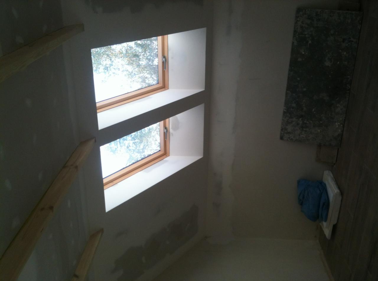 Dwa okna dachowe w surowym stanie, widoczne fragmenty konstrukcji drewnianej, ściany przygotowane pod malowanie z widocznymi śladami po szpachlowaniu, w rogu pomieszczenia leży niebieska szmatka.