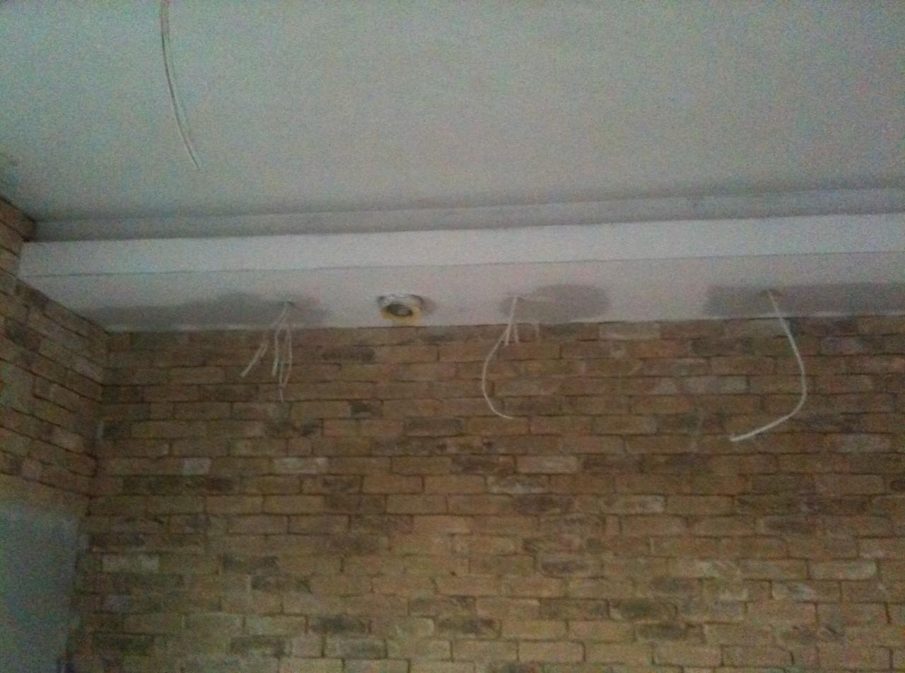 Częściowo wykończone wnętrze z ceglaną ścianą, podwieszonym sufitem z otworami na oświetlenie i wystającymi przewodami elektrycznymi.