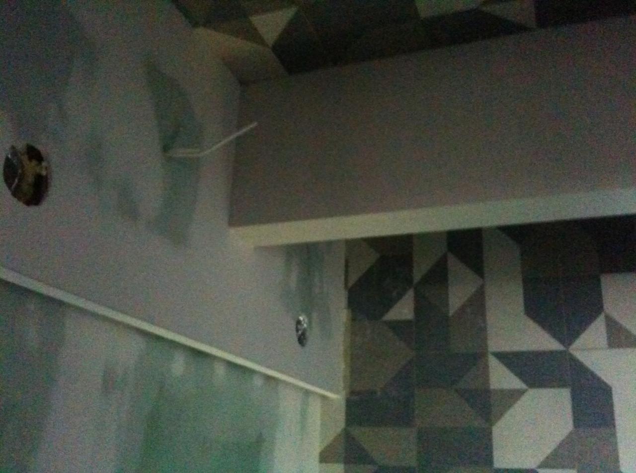 Fragment ściany z otworem na gniazdko elektryczne i wystającymi kablami, widoczna listwa przypodłogowa, ściana częściowo pokryta wzorzystymi płytkami.