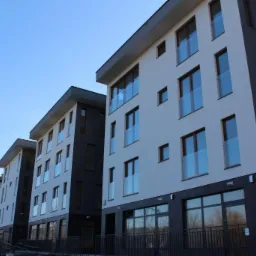 Nowoczesny budynek wielorodzinny z elewacją w kolorze białym i grafitowym, z balkonami z szklanymi balustradami, widok z dołu na tle błękitnego nieba.