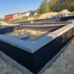 Zabezpieczone fundamenty budynku z izolacją przeciwwilgociową, widoczne niebieskie rury instalacyjne wewnątrz wykopu, w tle domy jednorodzinne i drzewa.