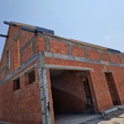 Wznoszenie ścian budynku z czerwonej cegły z widocznymi otworami okiennymi i drzwiowymi, betonowym wieńcem oraz przygotowaniem pod konstrukcję dachu, na tle błękitnego nieba.