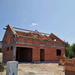 Surowy stan domu z czerwonej cegły z widocznym drewnianym szkieletem dachu, zbiornikiem na wodę i stertą cegieł na placu budowy w słoneczny dzień.