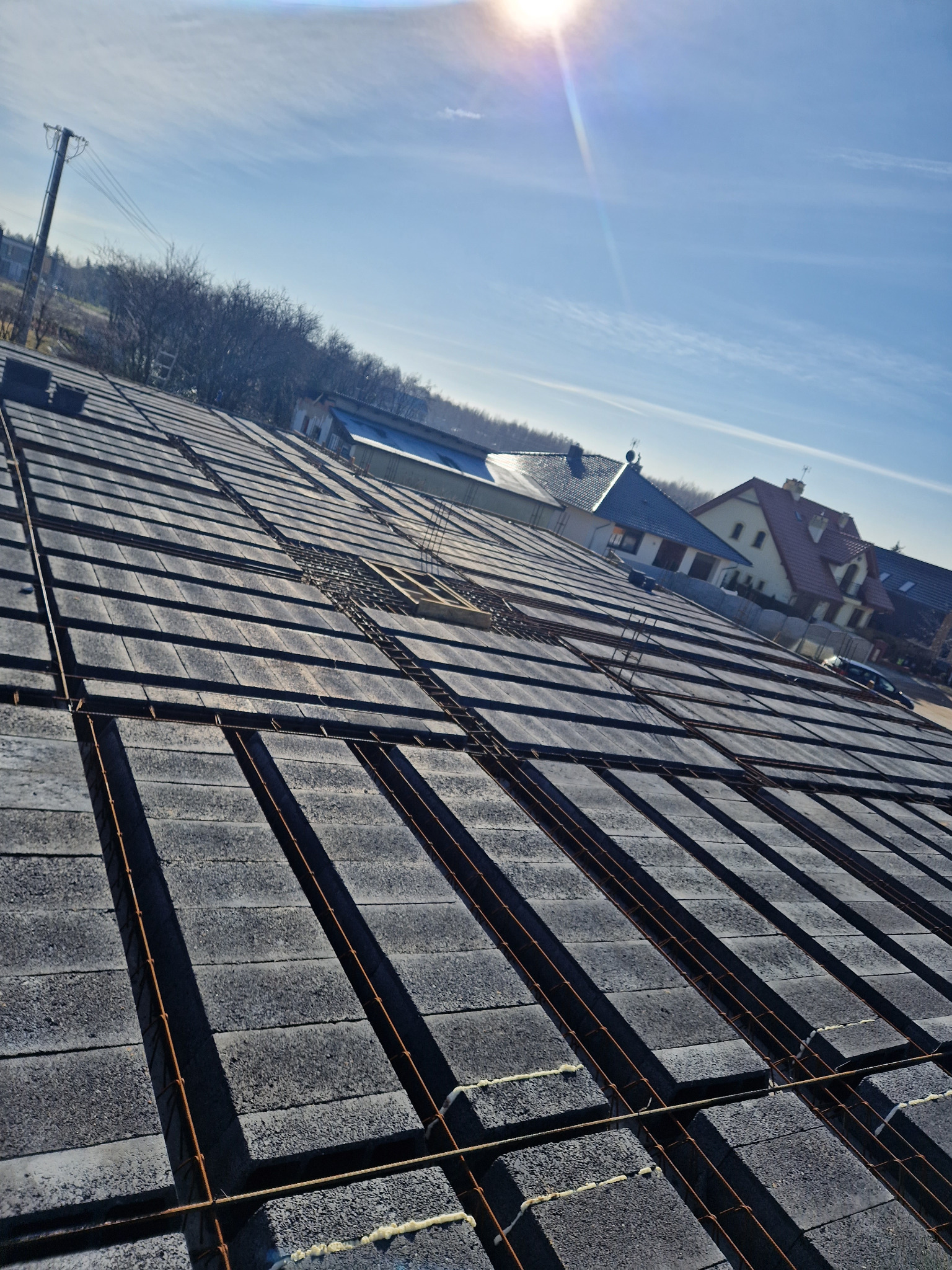 Wieniec stropowy Teriva w trakcie budowy domu jednorodzinnego, widok z góry na ułożone bloczki i zbrojenie, w tle zabudowa mieszkalna i błękitne niebo z promieniami słońca.