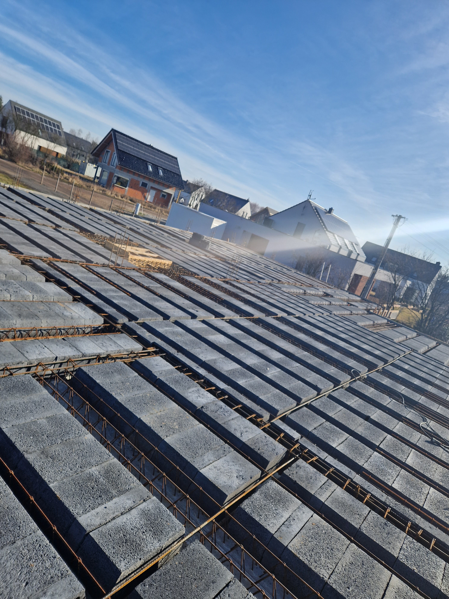 Wieniec stropowy z szalunków traconych i zbrojenia na budowie domu jednorodzinnego, widok z góry, w tle zabudowa mieszkalna.