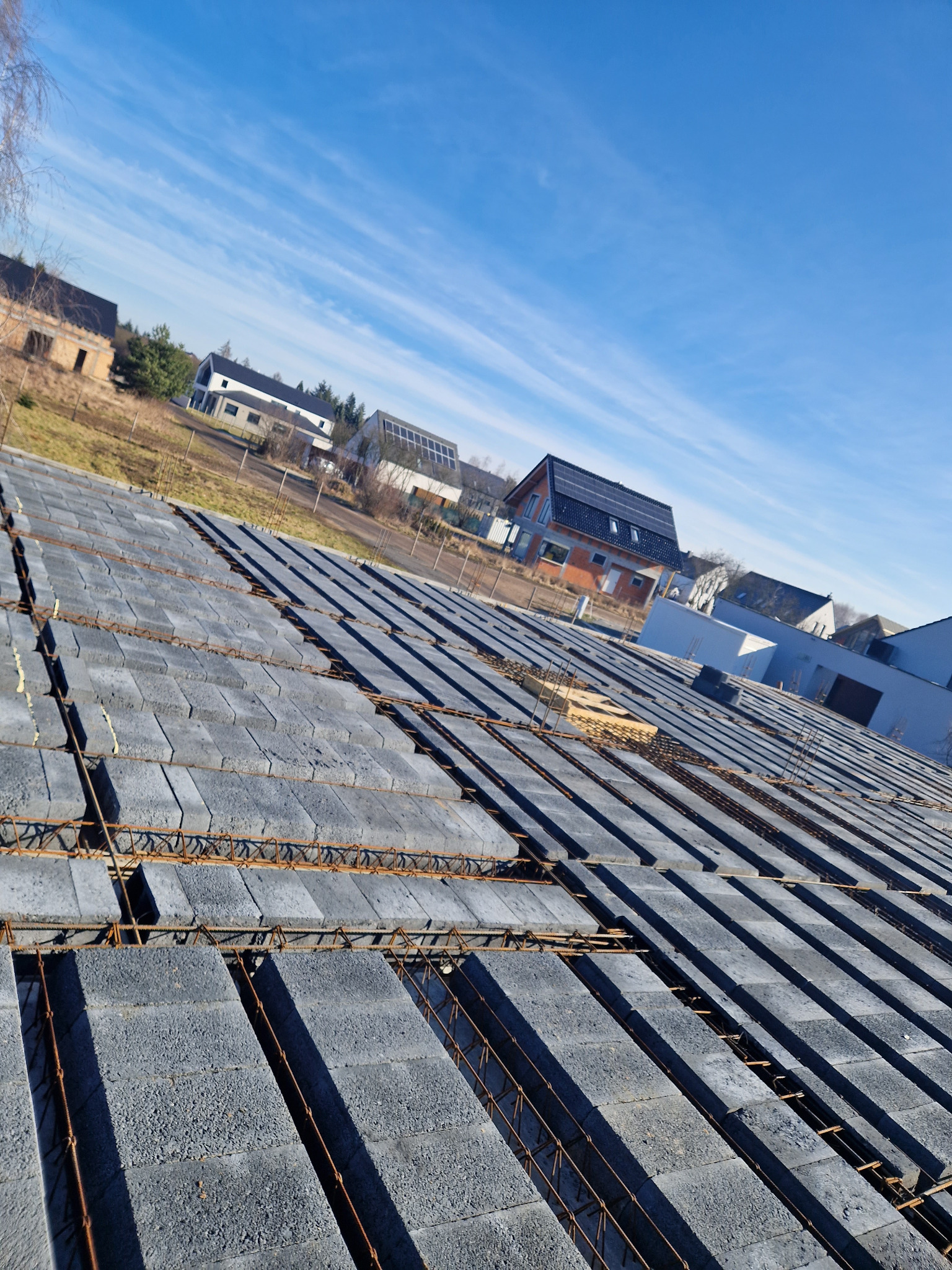 Wieniec stropowy z szarego betonu komórkowego zbrojony stalowymi prętami, w tle budynki mieszkalne w różnej fazie budowy i błękitne niebo.