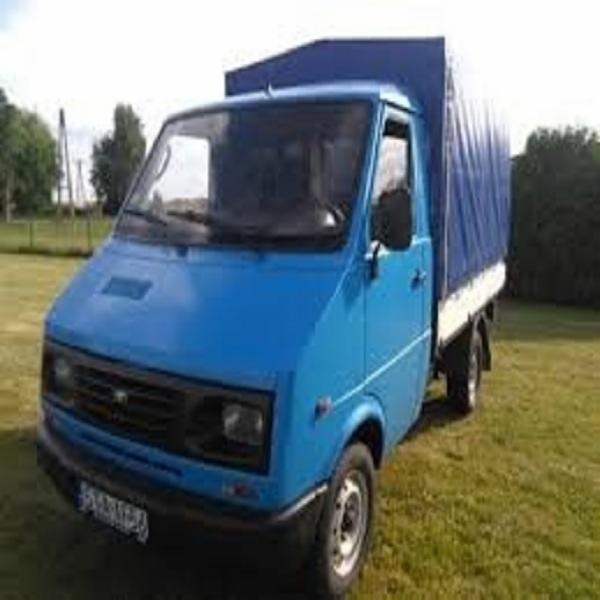 Niebieski samochód dostawczy z plandeką na trawiastym terenie, widok z przodu, lekko z boku.