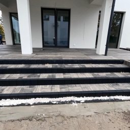 AS.PROJEKT Szymon Majewski - Schody wejściowe do nowoczesnego domu, wyłożone szarą kostką brukową z czarnymi obrzeżami, w trakcie prac wykończeniowych, widoczny piasek i biały grys.