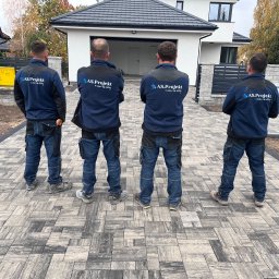 AS.PROJEKT Szymon Majewski - Ułożona kostka brukowa w odcieniach szarości przed nowoczesnym domem; czwórka pracowników firmy w granatowych bluzach z logo stoi tyłem, obserwując efekt końcowy.