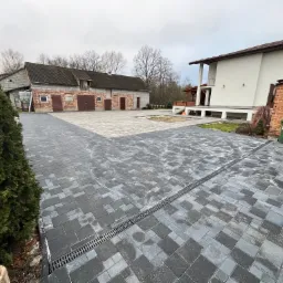 Nowo ułożona, szara kostka brukowa na podjeździe, z widocznym odpływem liniowym, domem z białym tynkiem i budynkiem gospodarczym w tle, w pochmurny dzień.