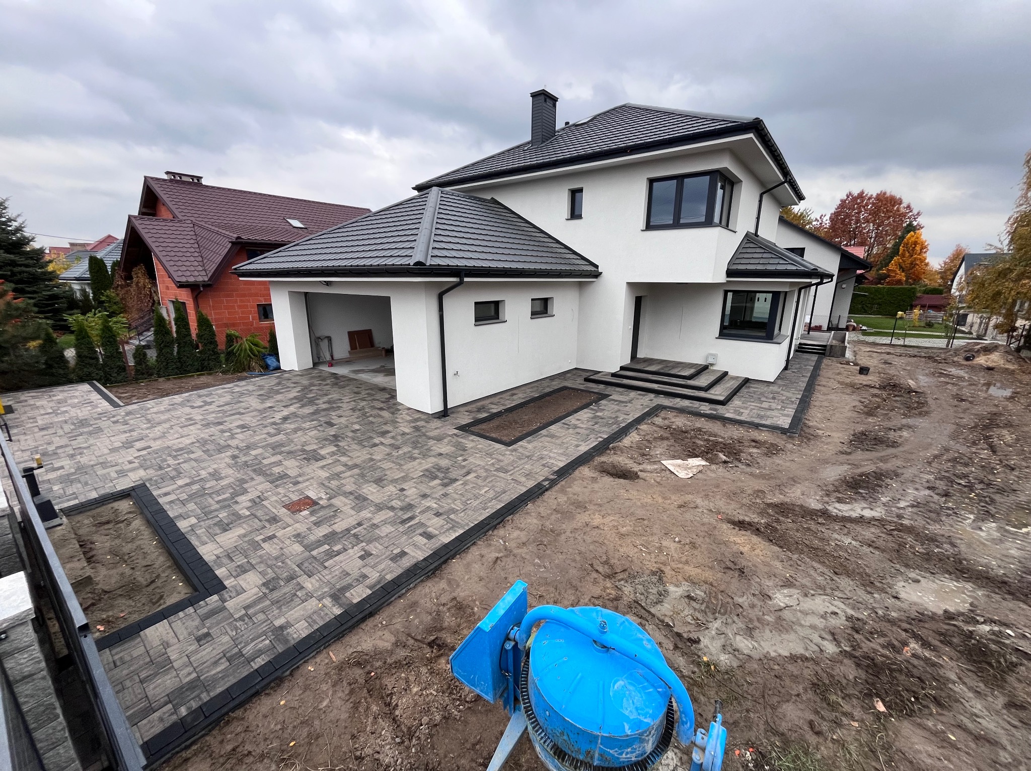 Nowoczesny dom jednorodzinny z szarą kostką brukową na podjeździe i wokół budynku, widoczna niebieska betoniarka w trakcie prac brukarskich.