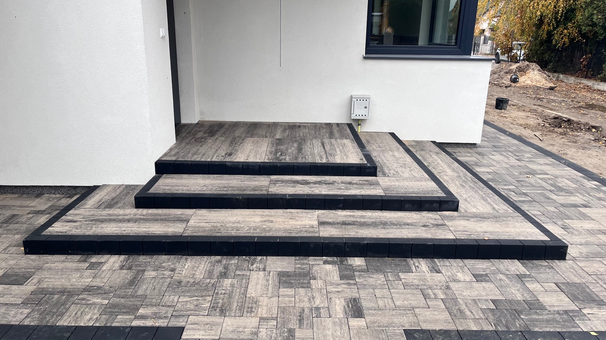 Nowoczesne schody wejściowe wyłożone szarą kostką brukową z czarnym obramowaniem, prowadzące do białego budynku z widoczną skrzynką gazową i fragmentem ogrodu z drzewami w tle.