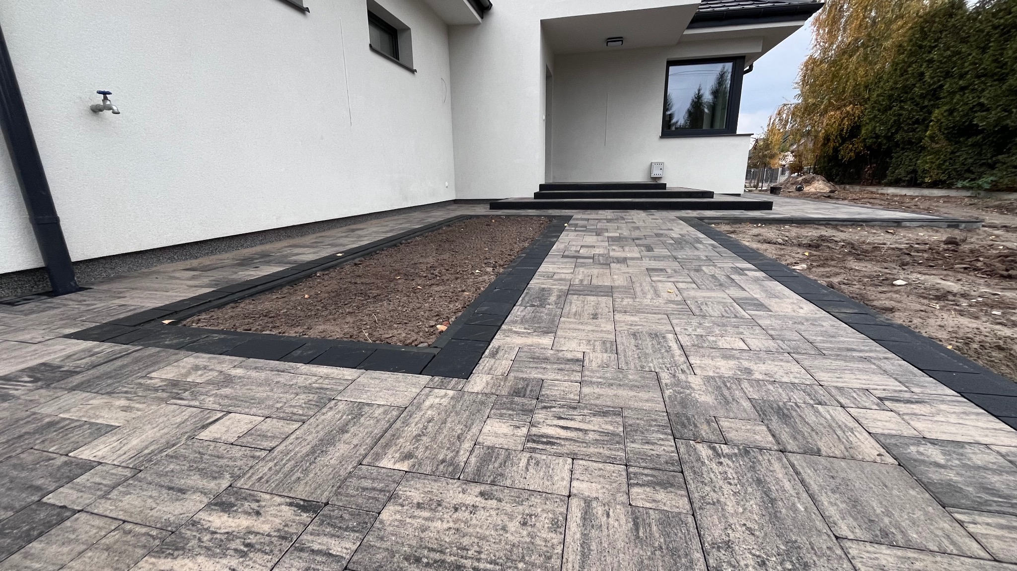 Ułożona szara kostka brukowa z czarnym obramowaniem rabaty przed nowoczesnym, białym domem z ciemnymi oknami i schodami wejściowymi.