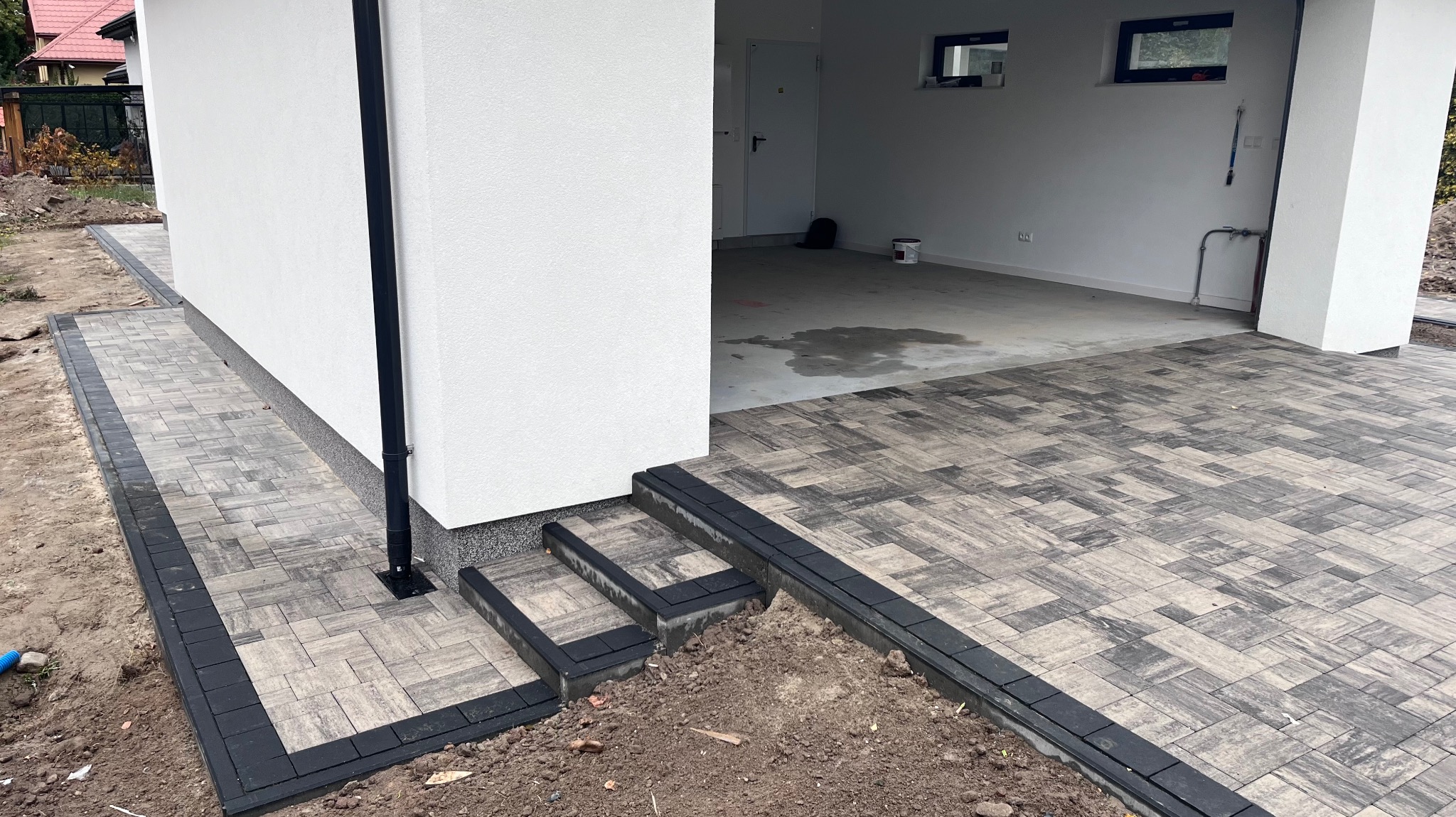 Ułożona kostka brukowa w odcieniach szarości tworząca ścieżkę i schody przed budynkiem z białymi ścianami, widoczny fragment rynny spustowej i wnętrze garażu.