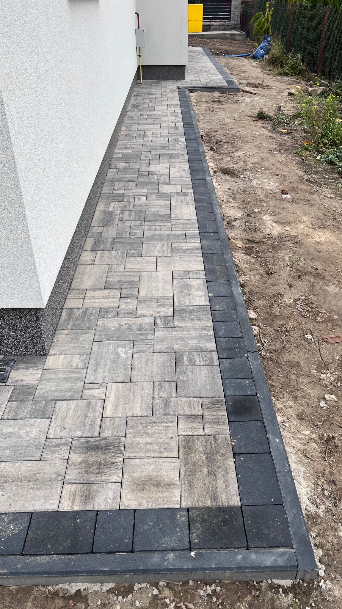 Świeżo ułożony chodnik z kostki brukowej w odcieniach szarości, obramowany ciemnymi kostkami, przylegający do białej ściany budynku i niezagospodarowanego terenu.