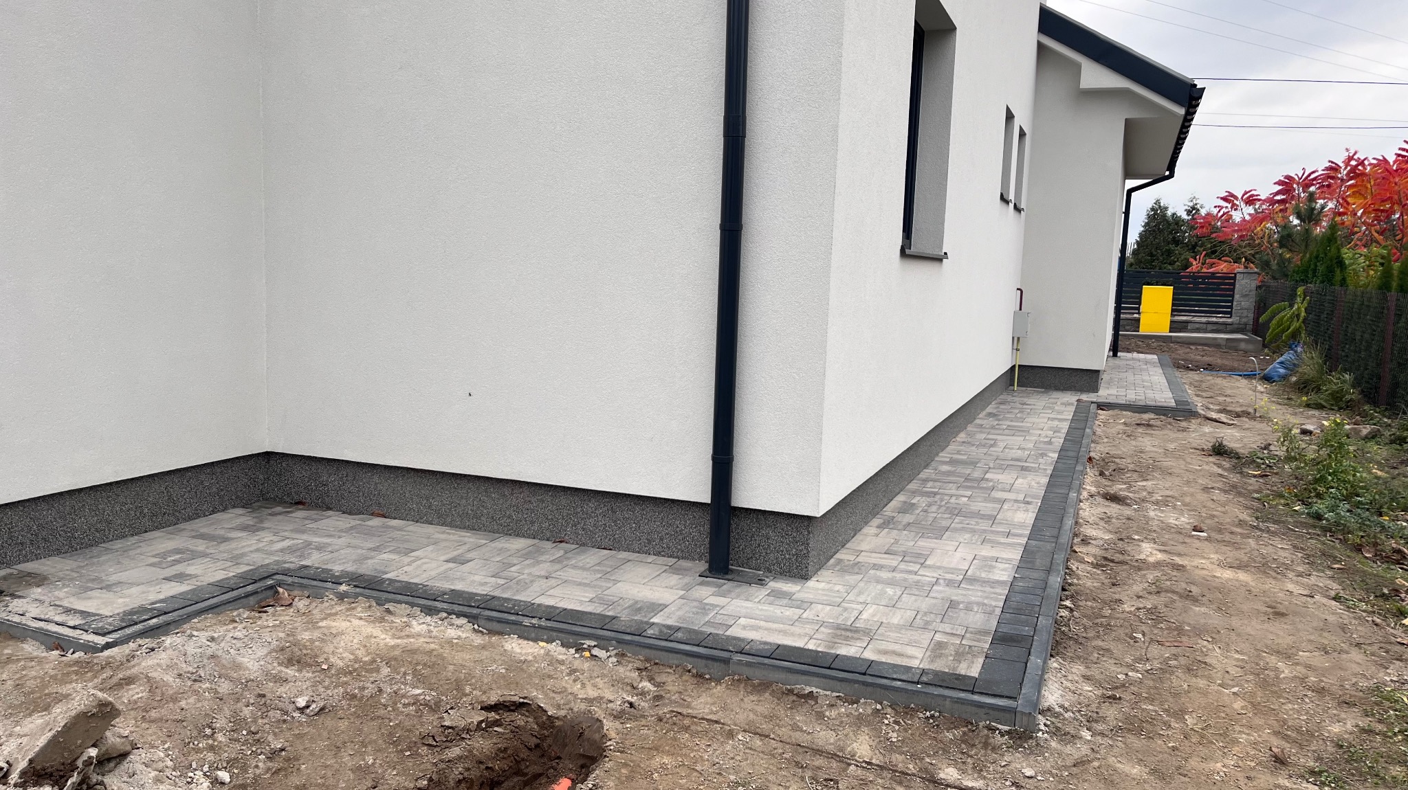 Świeżo ułożona, szara kostka brukowa tworząca chodnik wokół narożnika białego domu z czarną rynną, widoczny fragment wykopu pod dalsze prace.