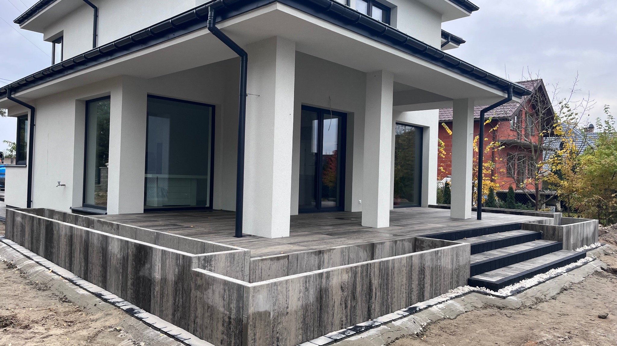 Nowoczesny taras z drewnianą podłogą i betonowymi elementami wykończeniowymi, widoczne schody z kostki brukowej i obrzeża z białym grysem.