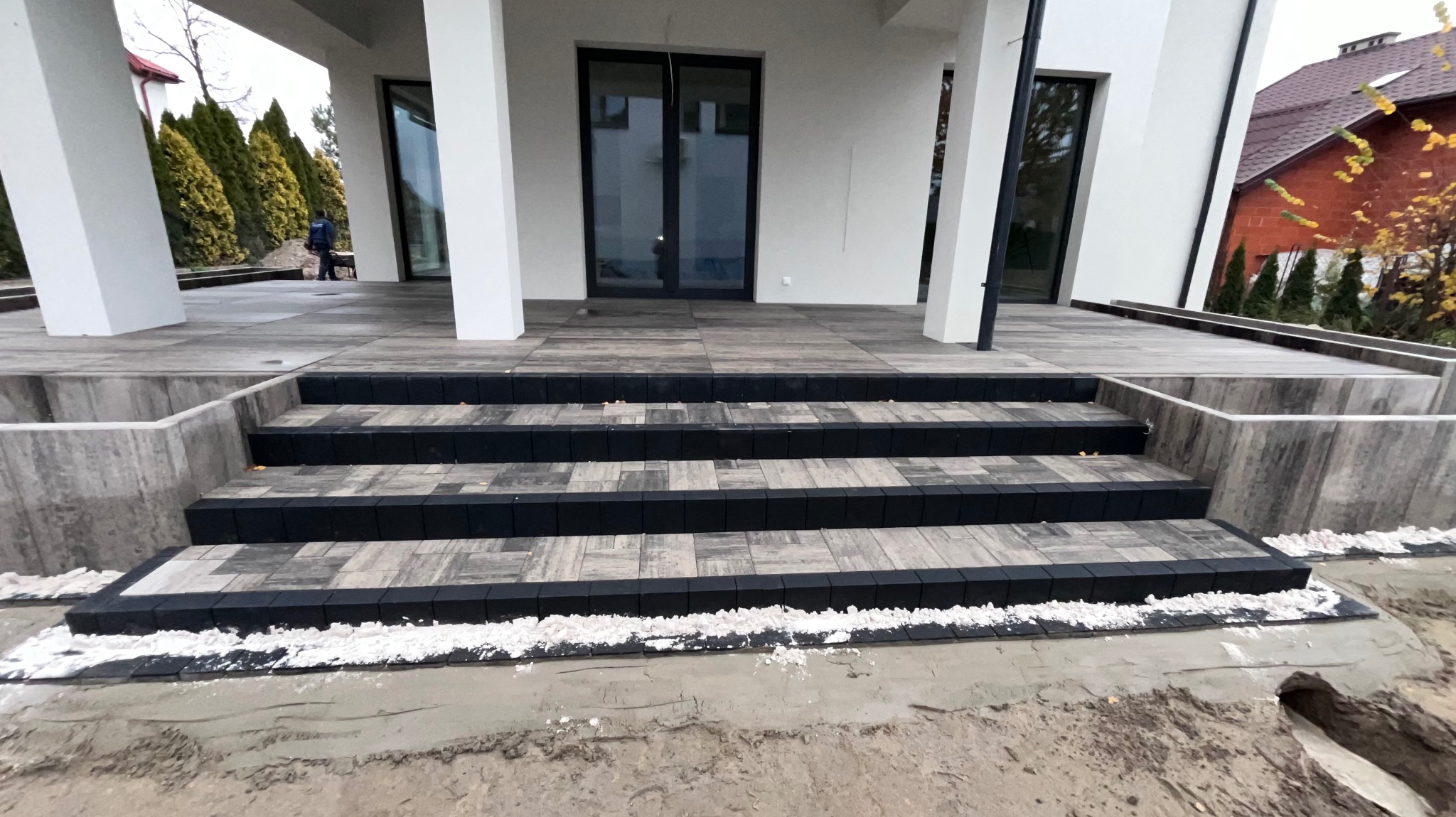 Schody wejściowe do nowoczesnego domu, wyłożone szarą kostką brukową z czarnymi obrzeżami, w trakcie prac wykończeniowych, widoczny piasek i biały grys.