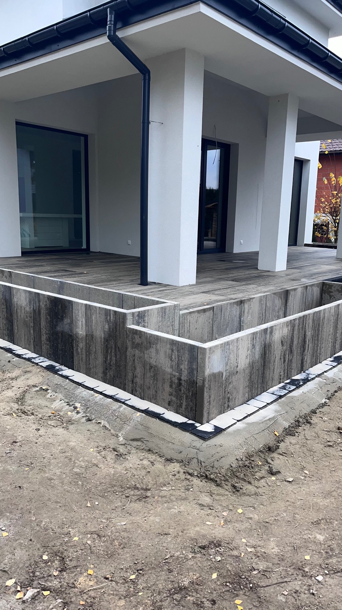 Fundament pod taras z betonowych elementów prefabrykowanych, widoczne czarne hydroizolacje na krawędziach, w tle nowoczesny dom z dużymi oknami.