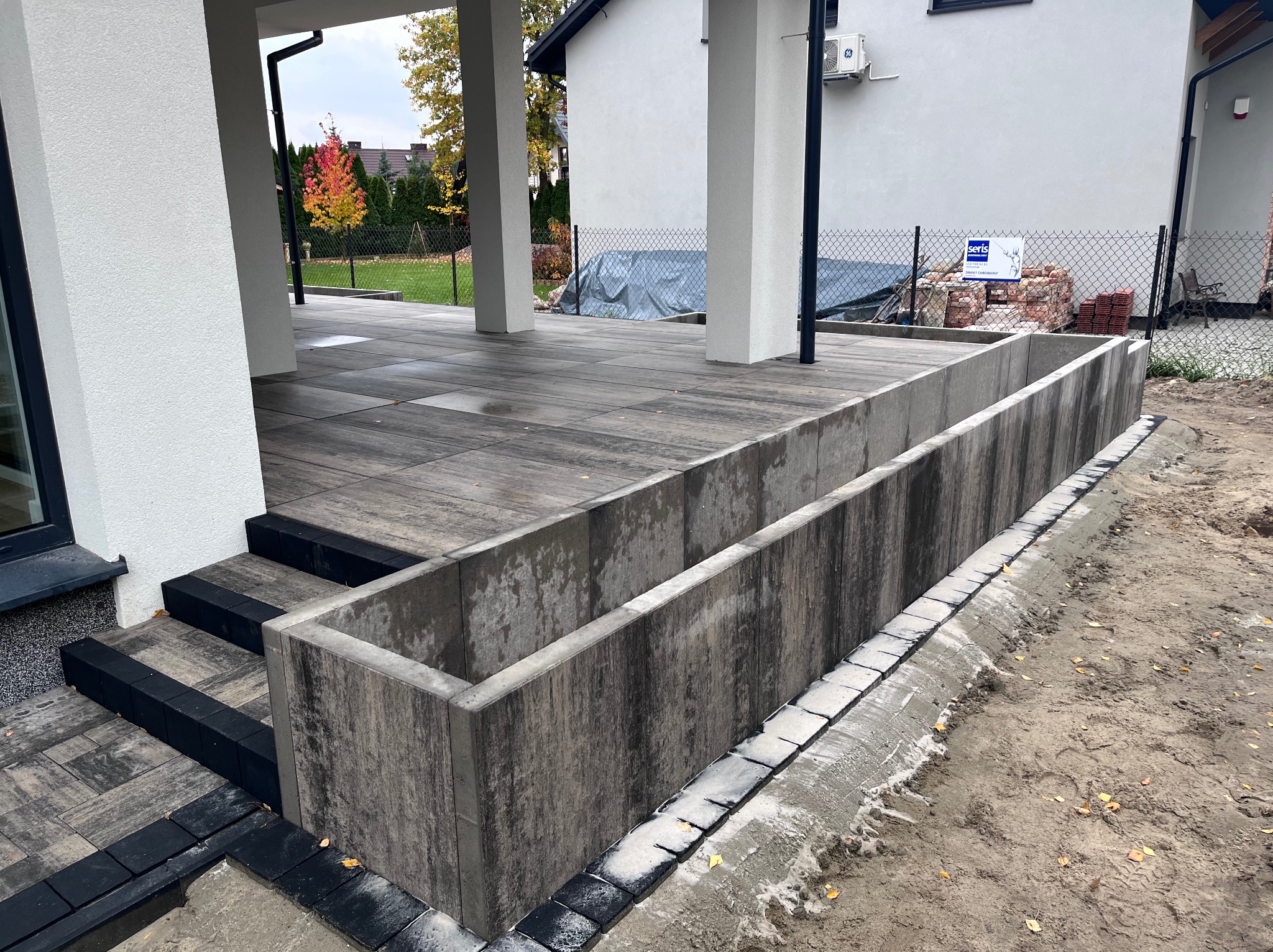 Nowoczesny taras z szarej kostki brukowej, schody z czarnymi obrzeżami, betonowa donica w surowym stylu, widok na ogród i dom.