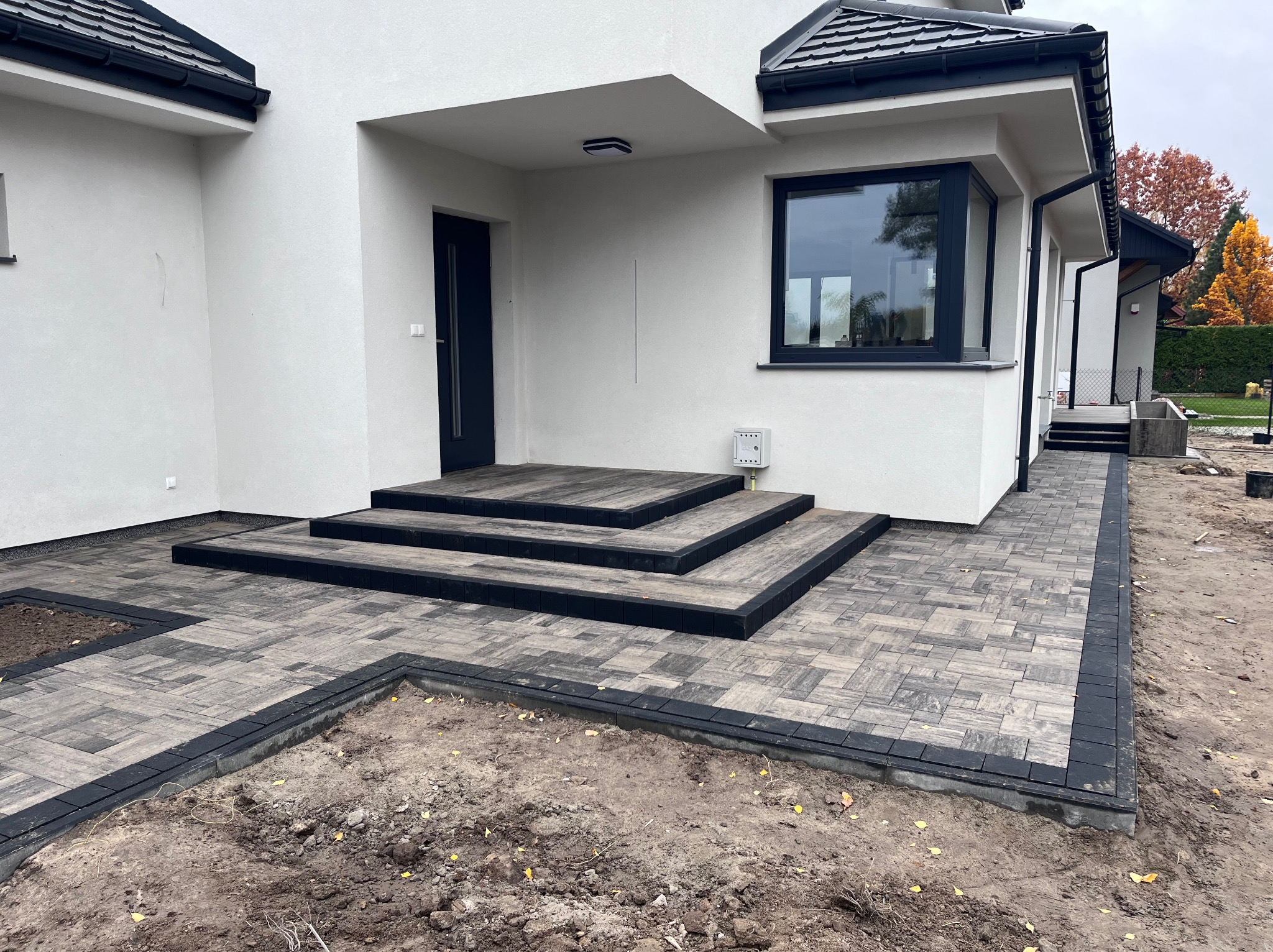 Nowoczesny podjazd i schody wejściowe z szarej kostki brukowej z czarnym obramowaniem, widoczne przygotowane rabaty ziemne.