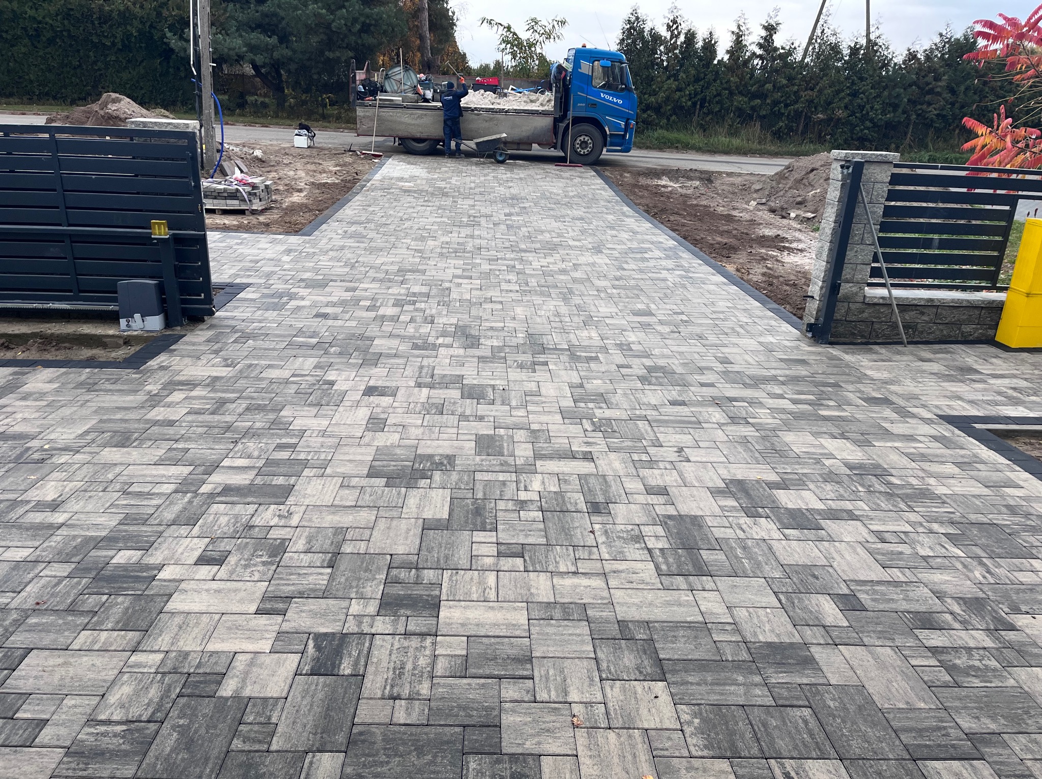 Nowo ułożona, szara kostka brukowa na podjeździe, z widocznym fragmentem bramy i samochodem dostawczym w tle.